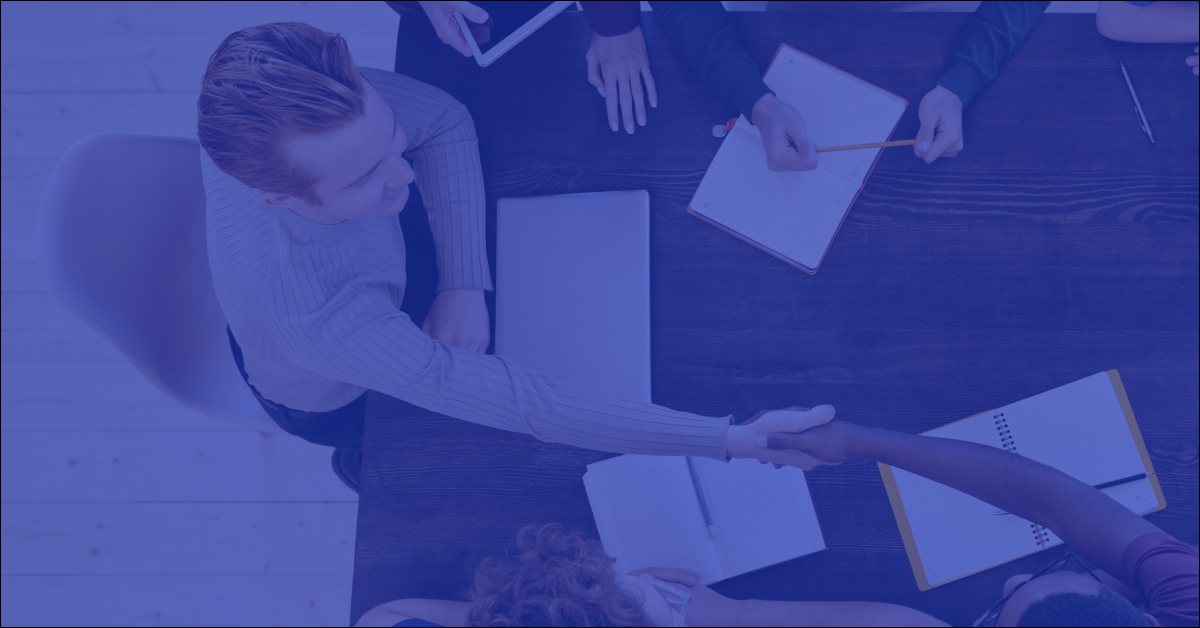 An overhead view of two professionals shaking hands across a dark conference table during a meeting, overlaid with a dark blue color filter.